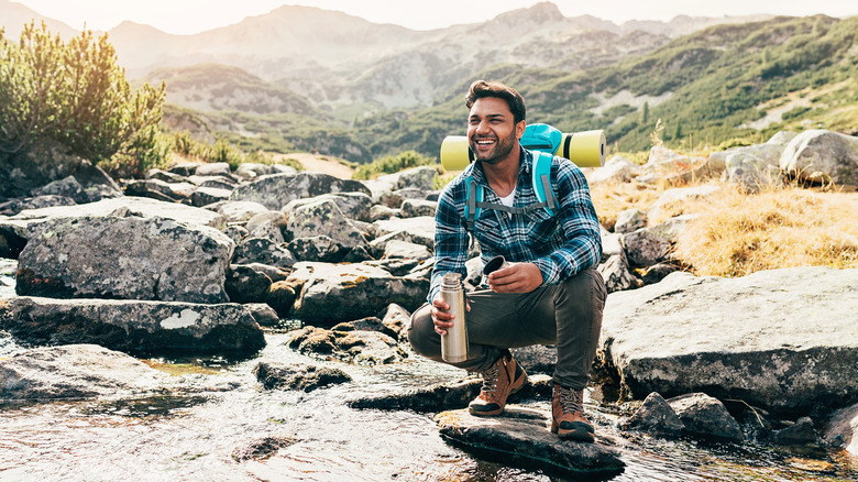 Happy hiker, crouched, holding water bottle over river