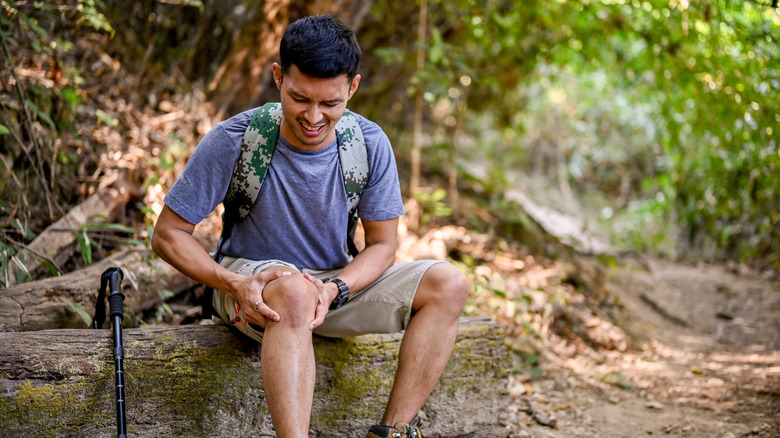 Male hiker sitting on log, injured and wincing, holding knee