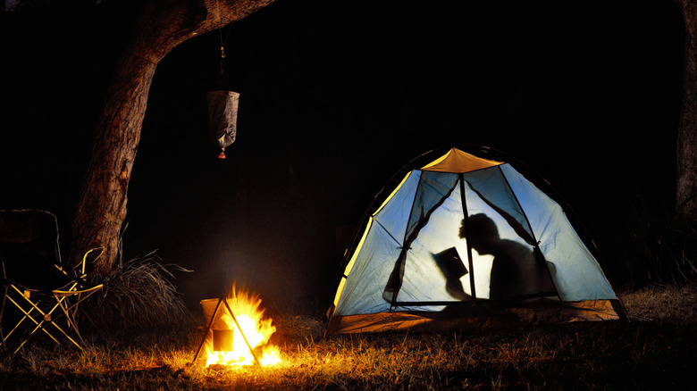 Person reading in tent at night, campfire outside