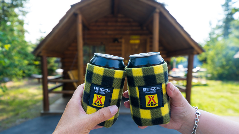 Two hands holding cans with foam sleeves in front of a tent