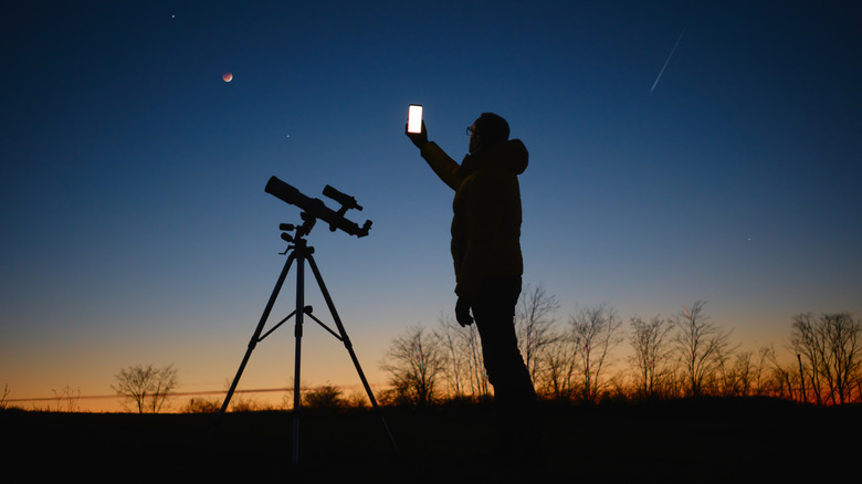 Silhouette of man standing next to telescope taking picture of night sky 